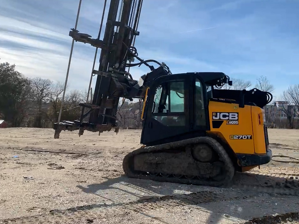 Side profile of a JCB skid steer rig equipped with vertical injection masts, positioned on a residential construction site with clear dirt surface.